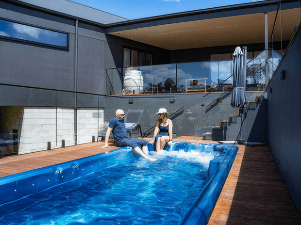 A modern residential courtyard with a narrow rectangular swimming pool surrounded by timber decking. Two people sit at the pool edge with their feet in the water. Dark rendered walls, extensive reflective glazing, an upper-level terrace, exterior stairs, and a covered outdoor area create a private, contemporary architectural setting.
