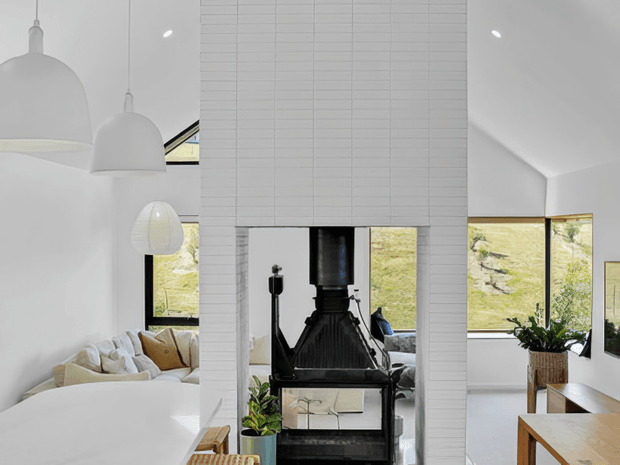 Bright, open-plan contemporary living and dining space with a dramatic white tiled chimney breast and black wood-burning stove as the central feature. Large windows frame a leafy outdoor view, while white pendant lights, pale upholstery, timber furniture and indoor plants create a clean, airy interior.