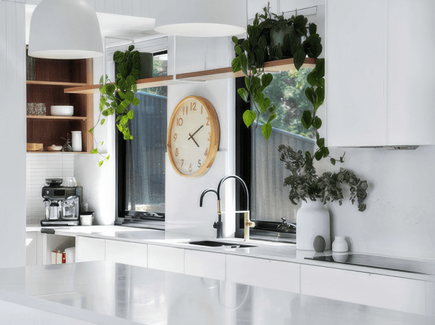 Bright, predominantly white contemporary kitchen with a large glossy island in the foreground, white flat-panel cabinetry, a white tiled backsplash, dark-framed windows, and a double sink with a high-arched faucet. Open timber shelving displays crockery and a coffee machine, while hanging pendant lights, a large round wall clock, and trailing potted plants add warmth and character.