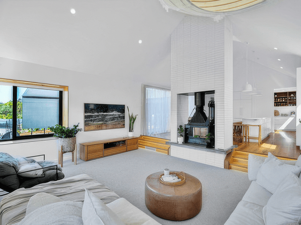 Bright, spacious open-plan living room with white walls and ceiling, pale carpet, large soft sofas, a round timber coffee table, timber media console, and a prominent white brick fireplace chimney with black firebox. Large windows and glazed doors bring in natural light, while timber steps lead toward a dining and kitchen area. The room has a refined contemporary coastal feel with warm timber accents.