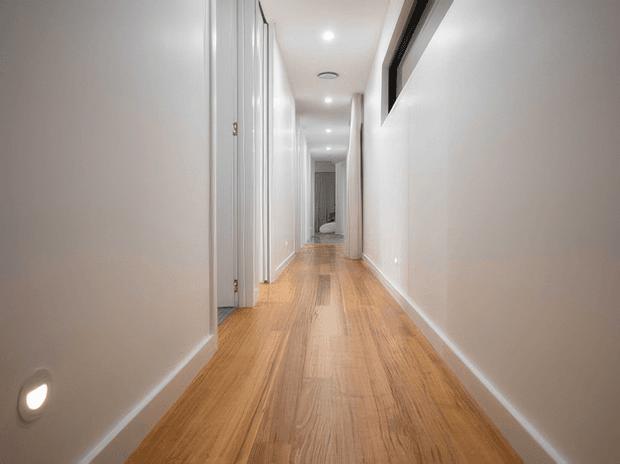 A long, narrow residential hallway with warm timber flooring, white walls and trim, multiple white doorways, recessed ceiling downlights, and a low wall light near the foreground. A dark-framed internal opening is visible high on the right wall.