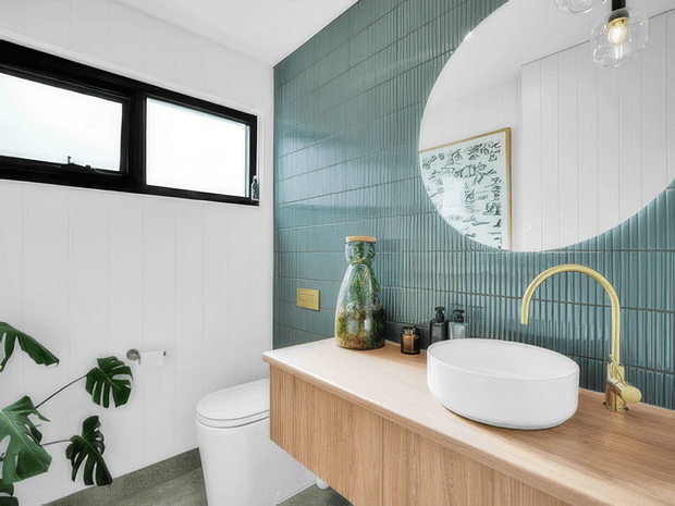 Bright contemporary bathroom featuring a floating timber vanity with a white vessel basin, brushed brass gooseneck tap, large round mirror, and full-height teal vertically ridged wall tiles. White vertical wall panelling, black-framed awning windows, a streamlined toilet, terrazzo-like grey flooring, and indoor greenery complete the fresh, modern scheme.