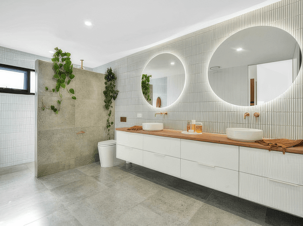 Bright contemporary bathroom featuring a long double vanity with white flat-front cabinetry, timber benchtop, two vessel basins and large round mirrors. The walls are clad in vertically ribbed white tiles, with a textured stone-look partition, concrete-look floor tiles, integrated greenery and a walk-in shower area.