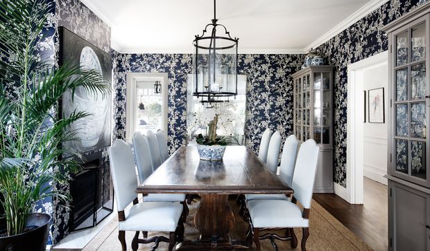 A formal dining room with a long dark wood dining table, upholstered dining chairs, ornate navy-and-white botanical wallpaper, a large black lantern pendant, traditional cabinetry, framed artwork, and abundant indoor palms. Glazed doors and openings connect to adjacent rooms, while white trim and ceiling balance the richly patterned walls.