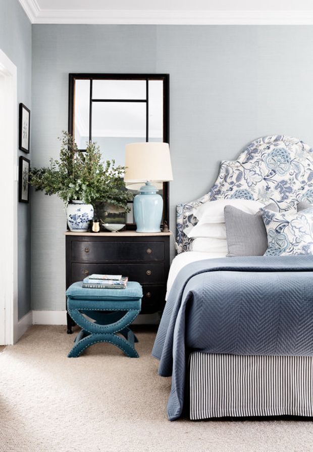 A refined bedroom corner with a blue upholstered bed, patterned blue-and-white bedding and headboard, a dark timber bedside chest, pale blue lamp, decorative mirror, framed artwork, and a small teal upholstered stool. The room has pale blue textured walls, white ceiling cornices and trim, and beige carpet.