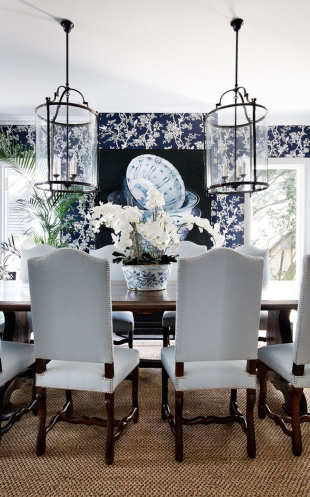 A refined dining room with a long dark wood dining table surrounded by upholstered white slipcovered chairs. The room features a bold navy-and-white botanical wallcovering, a dark fireplace or built-in feature wall with blue-and-white decorative plates, twin glass lantern pendant lights, a woven natural-fibre rug, and abundant white floral arrangements. Bright windows and greenery add an airy, elegant atmosphere.