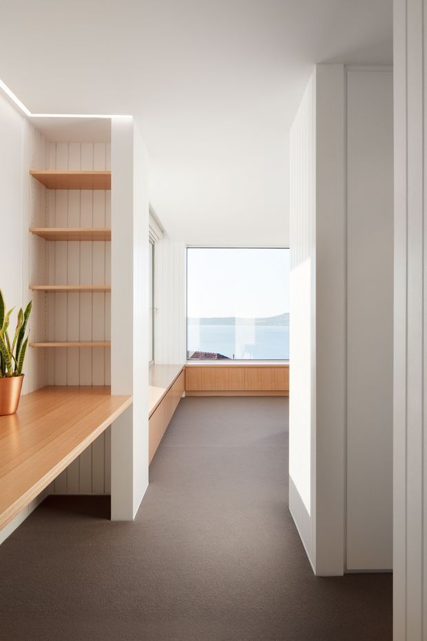 Bright minimalist interior corridor or study nook with extensive white vertical wall panelling, built-in light timber shelving and desk surfaces, and a large window overlooking water and hills. The space has a clean, calm feel with recessed linear lighting, integrated cabinetry, and a neutral carpeted floor.