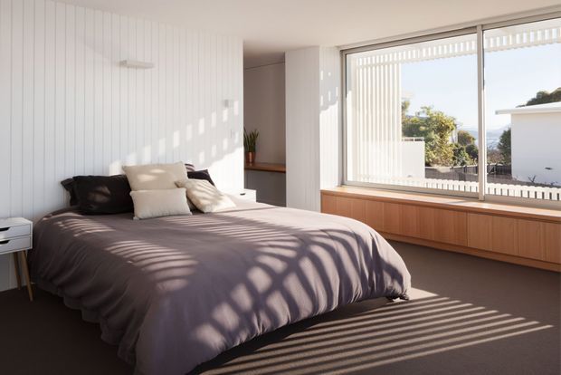 Bright, minimalist bedroom with a low upholstered bed dressed in muted mauve-grey bedding and layered neutral pillows. White vertical wall panelling, a large window with vertical blinds, and extensive built-in timber cabinetry beneath the window define the room. Strong sunlight creates striped shadows across the bed and carpet, while a leafy residential outlook is visible through the glazing.