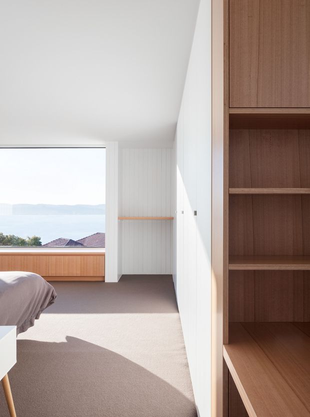 Bright, minimalist bedroom interior with a partial bed visible at left, a large picture window overlooking distant hills and rooftops, warm timber window-seat cabinetry, white vertical wall panelling, pale walls, and built-in dark timber shelving and wardrobe joinery along the right side. Soft neutral carpeting and strong sunlight create an airy, calm atmosphere.