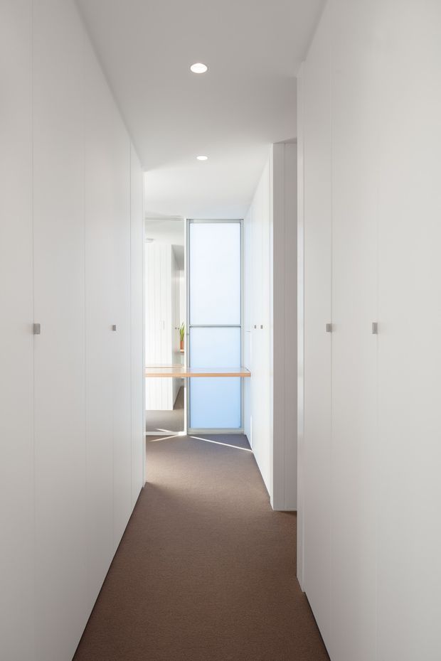 A narrow, bright minimalist hallway lined with full-height white built-in storage or cabinetry. The corridor has recessed ceiling downlights, a brown carpeted floor, and a frosted-glass door or window at the far end. A slim timber-toned surface projects across the hallway near the end, with a glimpse of an adjacent bright room beyond.