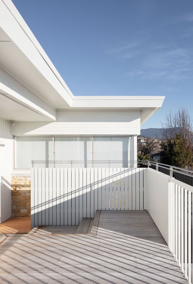 Bright exterior view of a modern white house with a flat roof, projecting eaves, white painted brick walls, large windows with horizontal blinds, and a raised timber deck or terrace enclosed by vertical white slat balustrades. Strong afternoon sunlight creates long linear shadows across the decking. A suburban hillside landscape is visible beyond the property.
