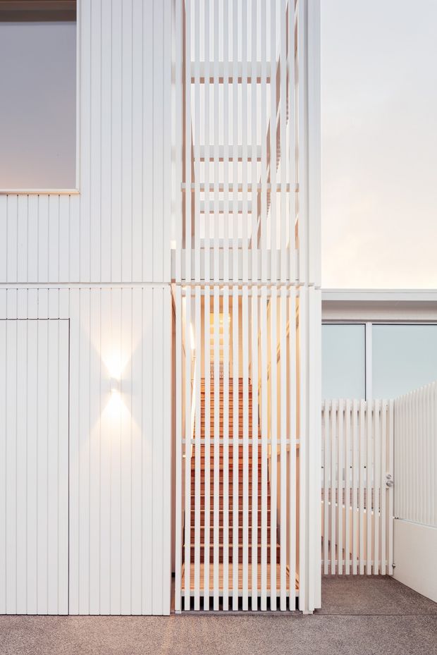 A bright, minimalist interior circulation space featuring a tall stairway screened by closely spaced vertical white slats. White vertical wall cladding, pale timber stair elements, warm integrated lighting, and a textured grey floor create a clean architectural composition. A glazed opening and matching slatted balustrade are visible to the right.