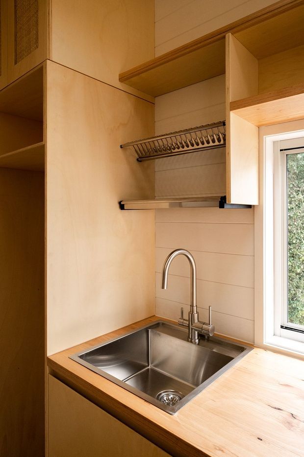 Compact kitchen sink area with a deep stainless-steel double-bowl sink, brushed metal gooseneck faucet, light timber benchtop, and extensive light plywood cabinetry. Open shelving and a hanging rack sit above the sink, with a white-framed window providing natural light. Horizontal timber wall panelling is visible behind the joinery.