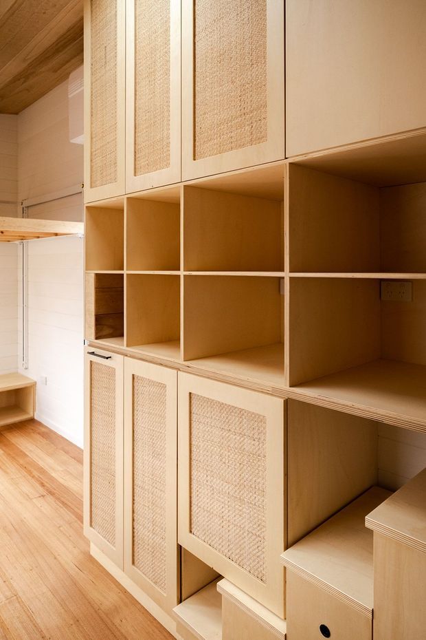 Built-in timber storage and cabinetry wall with open shelving, drawers, and framed doors featuring woven cane or rattan inserts. The warm plywood-toned joinery extends from floor to ceiling, set against white painted wall panelling and a timber floor. The space appears to be a compact entry or utility storage area with carefully integrated custom joinery.