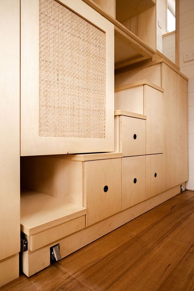 Close-up interior view of custom pale timber plywood built-in joinery, featuring stepped open shelving, multiple cabinet doors with black round pulls, a large inset woven cane panel, and concealed storage integrated into a wall. Warm timber flooring and partial white wall panelling are visible.