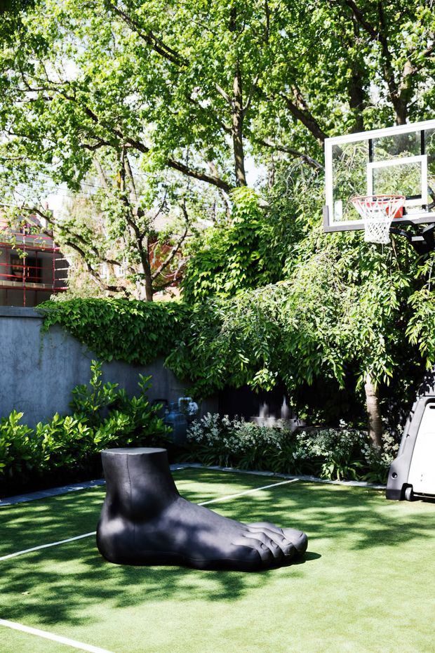 A leafy outdoor recreational area with an artificial grass basketball court, white court markings, a basketball hoop, and a large black novelty foot-shaped sculpture or seat. Dense mature planting, hedges, and a rendered boundary wall surround the space.