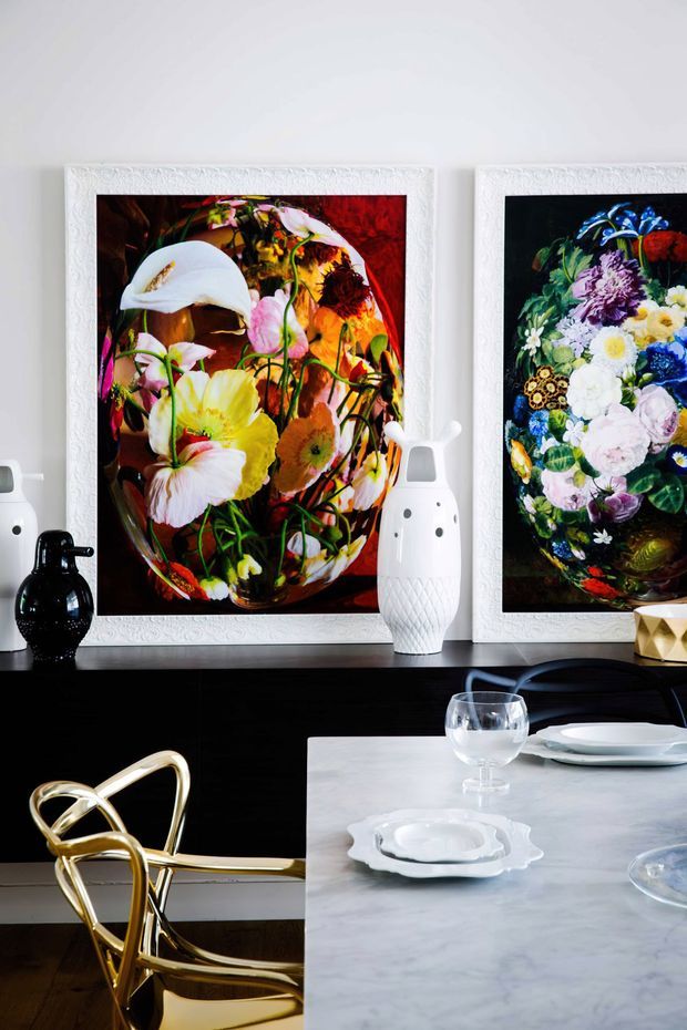 A richly styled contemporary dining area with a glossy black sideboard, colourful floral artwork in ornate white frames, sculptural vases, and a pale stone dining table set with white dishes and glassware. A reflective gold sculptural dining chair adds a bold metallic accent.