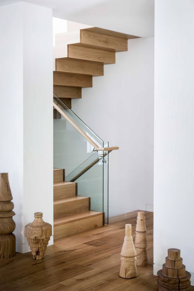 Bright minimalist interior hallway featuring a sculptural timber staircase with cantilevered oak treads, clear glass balustrade, pale timber handrail, white walls, and matching timber flooring. Decorative carved wooden objects are arranged on the floor around the stair.