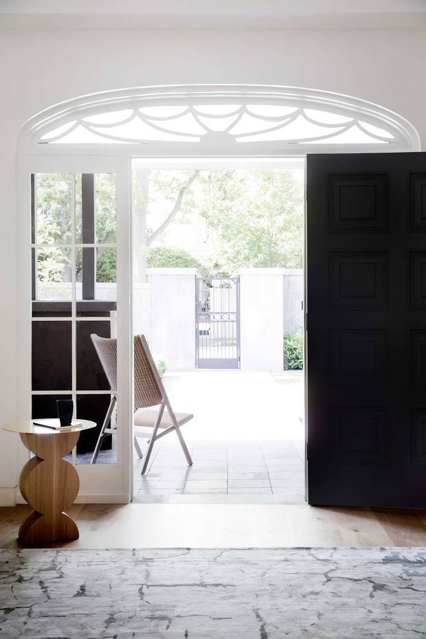 Bright, elegant entryway viewed from inside, featuring a large dark paneled front door opened toward a light-filled courtyard or exterior area. The doorway has an ornate fanlight with curved decorative glazing details, while a glazed side panel and French-style internal door create additional transparency. A sculptural wooden side table, chair, and pale stone or timber flooring sit in the foreground, with a large grey patterned rug adding softness.