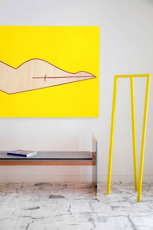 A minimalist interior vignette with a bright yellow abstract artwork above a slim console table. The console combines a dark speckled stone or terrazzo-like top, warm timber edging, and slender glass supports. A tall, angular yellow-framed mirror leans against the pale wall, while the floor has a distinctive pale, heavily veined stone finish. The composition is graphic and colour-blocked, with yellow accents contrasted against white, timber, and stone.