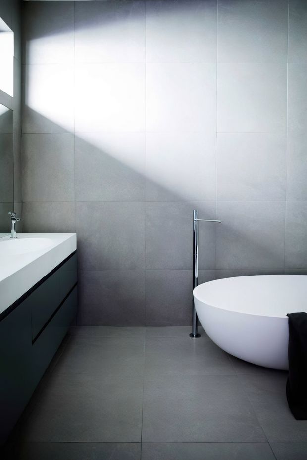 A minimalist contemporary bathroom featuring large-format grey wall and floor tiles, a dark floating vanity with a white benchtop, a freestanding white oval bathtub, and a slim chrome floor-mounted tap. Strong natural light creates a diagonal shadow across the tiled wall.
