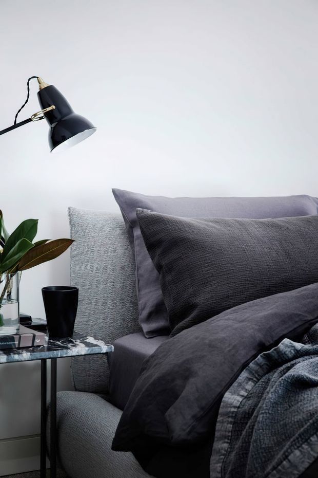 Close-up interior photograph of a styled bedroom corner featuring a grey upholstered bed, layered charcoal and grey bedding and pillows, a black wall-mounted reading lamp, and a small marble-topped side table with a plant and black cup. The palette is dark, cool, and refined against a light wall.
