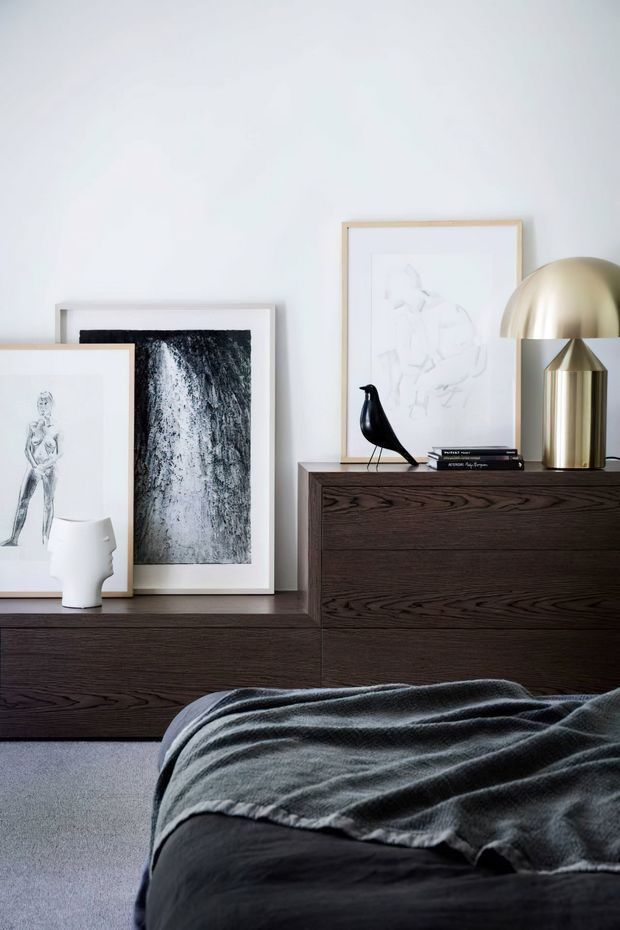 A moody, refined bedroom vignette featuring a dark timber dresser and low sideboard, layered framed monochrome artwork, sculptural white ceramic decor, a black bird ornament, and a brass dome table lamp. The foreground shows a dark upholstered bed draped with a charcoal grey throw, set against a soft white wall and grey carpet.