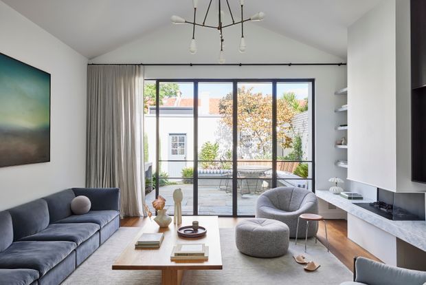 Bright contemporary living room with a pitched ceiling, full-height black-framed sliding glass doors opening to a courtyard, a deep blue sectional sofa, sculptural grey lounge chair and ottoman, timber coffee table, white built-in media cabinetry, and a linear fireplace with a pale stone surround. A modern multi-arm pendant hangs centrally, while open shelving and neutral curtains complete the refined interior.
