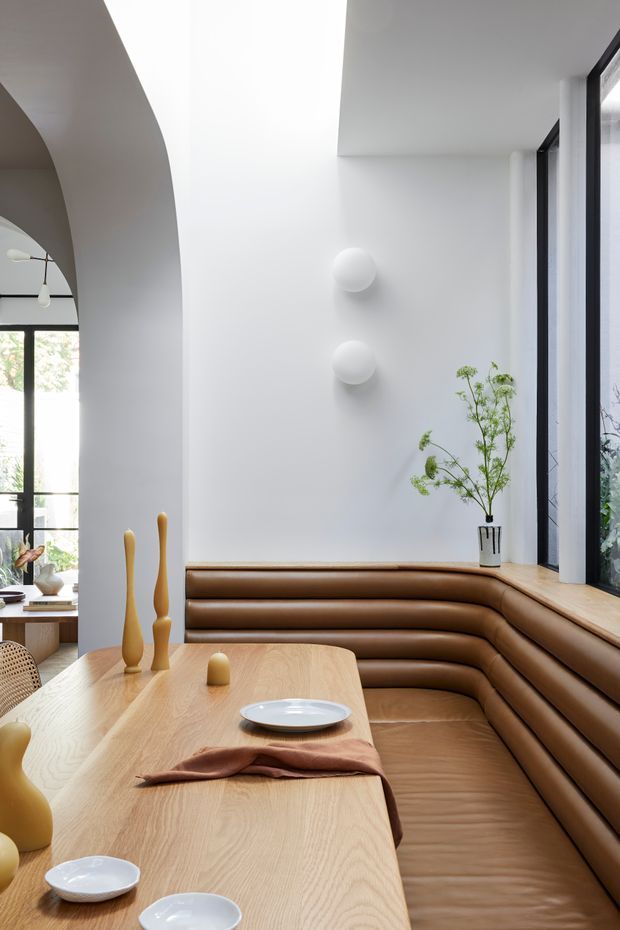 A bright, refined dining nook with a large built-in curved banquette upholstered in warm brown leather, paired with a pale timber dining table. The room features white sculptural walls, a broad arched opening, black-framed floor-to-ceiling glazing, simple globe wall lights, and restrained natural decor.