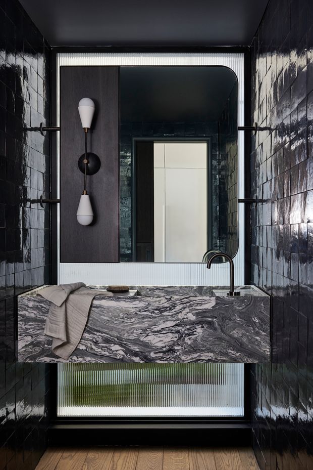 A dramatic compact bathroom with glossy dark wall tiles, a floating vanity clad in heavily veined grey-and-white stone, black fixtures, and a large rounded-corner mirror. Ribbed translucent glazing behind the vanity and mirror brings in filtered light, while a sculptural vertical wall sconce and timber floor add contrast.