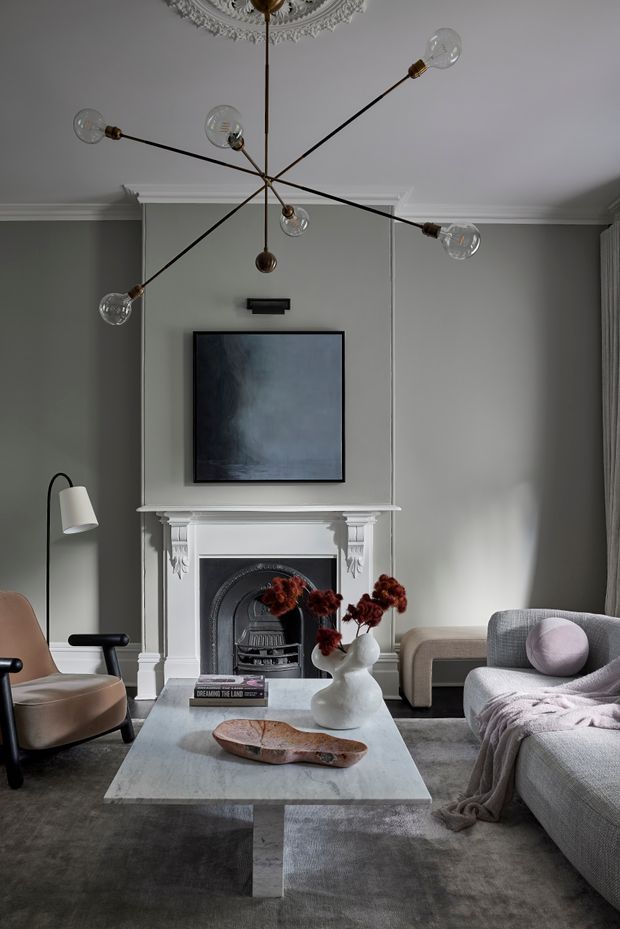 A refined living room with a decorative traditional fireplace, grey painted walls, ornate ceiling rose and cornices, a contemporary multi-arm pendant light, wall-mounted television, sculptural coffee table, upholstered armchair and sofa. The room combines classical architectural detailing with modern furniture and restrained neutral tones.