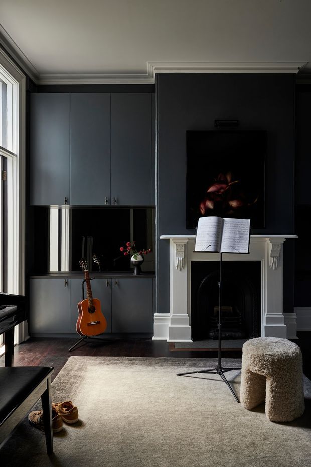 A moody, refined living room with dark blue-grey built-in cabinetry, a traditional white fireplace, and a wall-mounted television. A music stand with open sheet music stands in front of the fireplace, while a guitar, piano edge, sculptural boucle stool, and large neutral rug create a music-focused lounge setting. Tall windows bring in soft natural light, and decorative ceiling cornices add architectural detail.