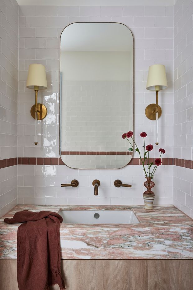 A refined bathroom vanity with a large rounded-rectangle mirror, glossy white subway wall tiles, and a horizontal terracotta-brown tile accent band. Brass wall sconces with cream shades flank the mirror above a wall-mounted brass tap set. The vanity has a dramatic pink, white, and grey stone benchtop with an integrated rectangular basin and light timber cabinetry below. A rust-coloured towel and a small vase of flowers add warmth.
