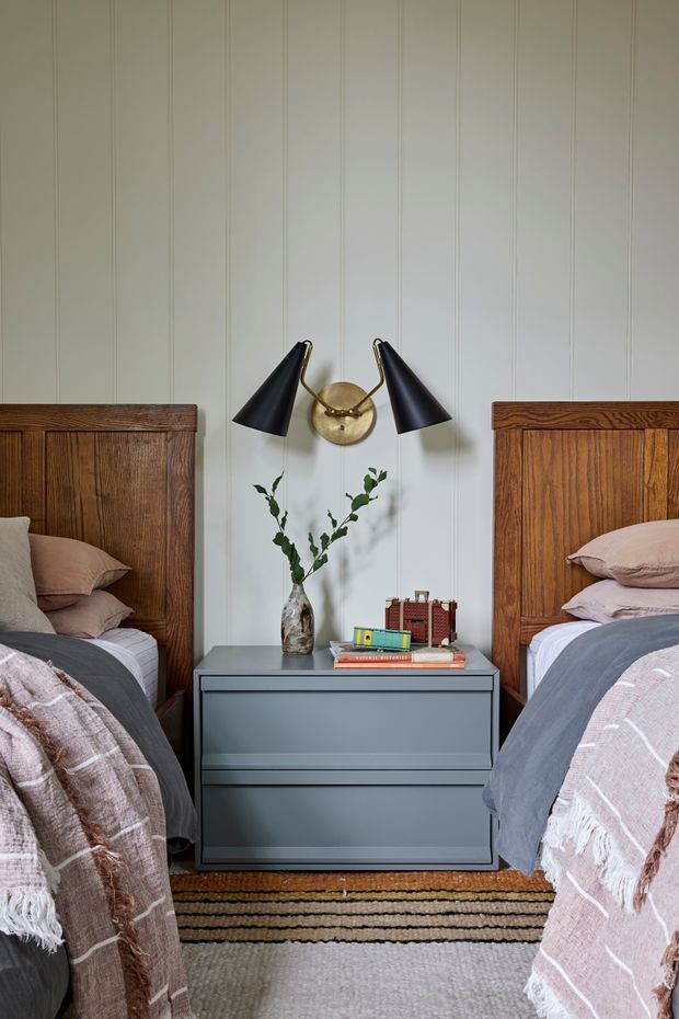 A symmetrical bedroom vignette with two twin beds flanking a dark blue-grey bedside cabinet. The room features warm carved timber headboards, pale vertical wall panelling, black-and-brass wall sconces, layered muted bedding, a small vase of greenery, books and decorative objects, and a striped woven rug.