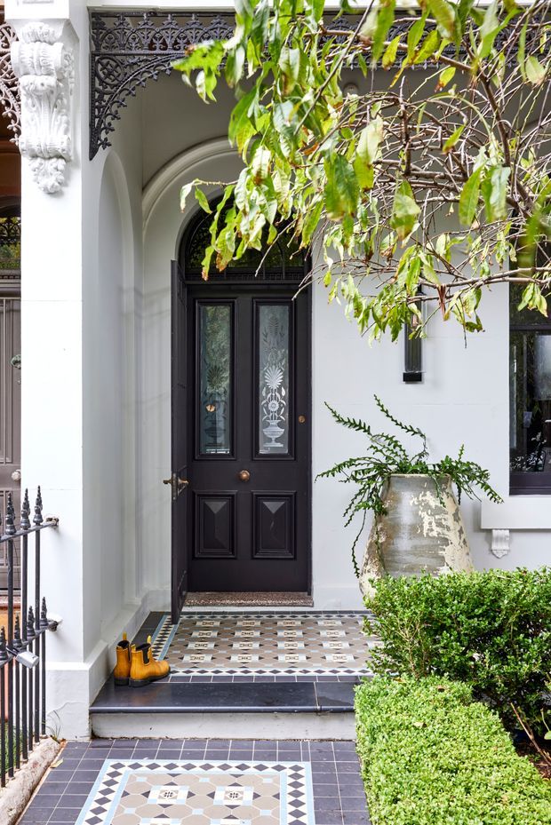 A refined traditional townhouse entrance with a black decorative front door set within a white rendered facade and arched entry surround. Ornate cast-iron detailing, carved architectural ornament, patterned encaustic-style tiled steps and path, dark stone thresholds, a large ceramic planter, and clipped greenery create a formal heritage character.