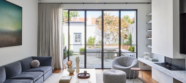 A bright contemporary living room with a grey sofa, rounded lounge chair and ottoman, low coffee table, and wall-mounted artwork. Full-height black-framed glazing and curtains open onto a paved courtyard or terrace, while a sleek white built-in media wall and shelving line the right side. The room has timber flooring, neutral finishes, and abundant natural light.