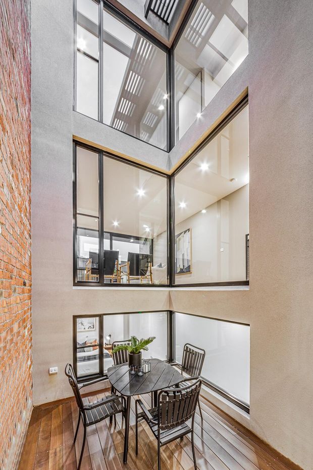 Tall multi-level interior atrium or void with a compact dining area in the foreground. The space features exposed red brick, textured plaster walls, extensive black-framed internal glazing, warm timber flooring, and a high ceiling with strong daylight and linear skylight or clerestory-like openings. Upper-level rooms and circulation spaces are visible through the glazing, creating a dramatic vertical connection between floors. A dark timber dining table with metal chairs occupies the lower level.