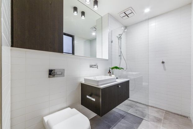 Bright contemporary bathroom with white rectangular wall tiles, a large mirrored vanity wall, dark wood-look floating vanity with white vessel basin, wall-mounted toilet, chrome fixtures, and a walk-in shower with clear glass. Grey-toned tiled flooring and recessed ceiling lighting complete the clean, modern fit-out.