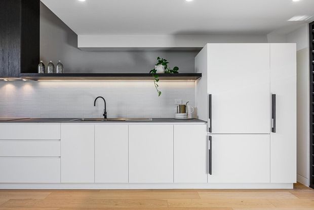 A contemporary minimalist kitchen with a long run of white flat-panel cabinetry, integrated tall storage, dark vertical handles, a dark benchtop, and a black gooseneck tap. A ribbed light backsplash is illuminated by concealed under-cabinet lighting, while dark timber-toned overhead cabinetry and open shelving create contrast. The kitchen opens onto a light timber floor and has a clean, streamlined appearance.