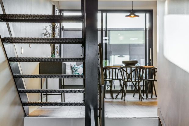 Interior view from a stairway, showing an open-riser staircase with dark metal stringers and textured treads, leading toward a bright dining area. The dining space includes a timber table, dark spindle-back chairs, pendant lighting, large glazed doors or windows, and a contemporary open-plan layout.