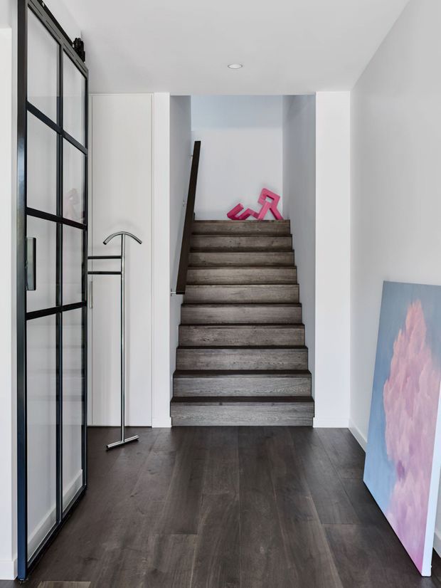 A minimalist residential stairway viewed from a dark-floored entry hall. The staircase has dark weathered timber treads and a simple dark handrail, set between white walls with recessed ceiling lighting. A black-framed glazed partition or door is visible on the left, with a tall chrome coat stand and a colourful abstract artwork leaning against the right wall.
