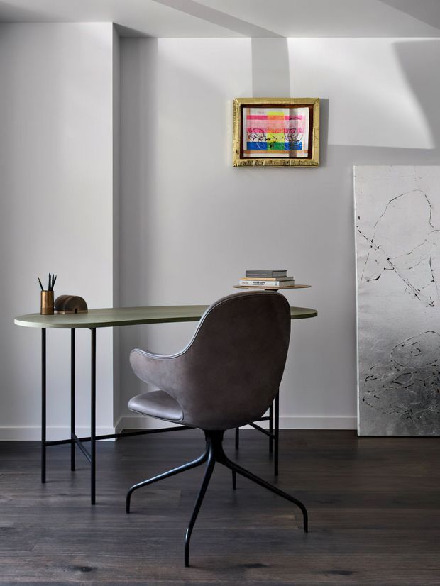 A refined minimalist home office or study with a slim greenish built-in or console-style desk, slender black metal legs, a sculptural upholstered swivel chair, dark timber flooring, and pale grey walls. Abstract artwork and a small framed colourful artwork add restrained visual accents, while daylight creates geometric shadows across the walls.