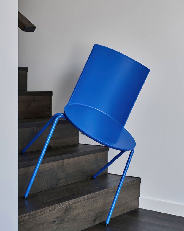 A vivid blue modern chair with a curved molded seat and back, slim tubular legs, and arm supports is positioned on dark timber stairs beside a plain white wall. The image highlights the chair as a contemporary furniture product within a stairway setting.