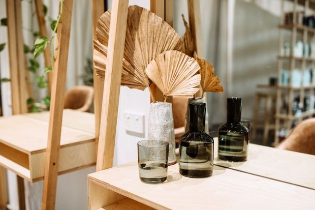 Warm, styled interior vignette featuring a light timber console or shelving unit with dark glass bottles, a textured ceramic vase, a drinking glass, and dried fan-shaped palm leaves. A large mirror reflects a timber dining table, chair, shelving, and indoor greenery, while vertical timber framing and light neutral finishes create a natural, contemporary feel.