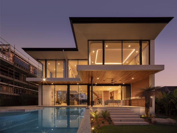 Twilight exterior view of a contemporary multi-level residence with strong horizontal cantilevered roof slabs, extensive floor-to-ceiling glazing, warm interior lighting, and timber-lined soffits. A swimming pool occupies the foreground, with landscaped planting and stepped access leading to the covered outdoor living area. The neighbouring structure is visible at left but is secondary.