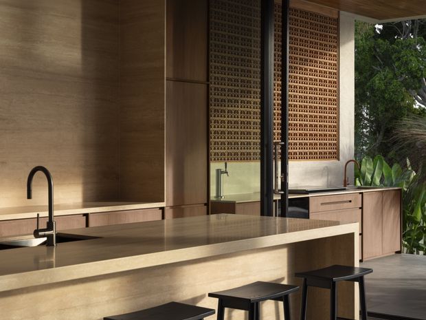A sophisticated open-plan kitchen with warm timber cabinetry and wall panelling, a large stone island with integrated sink and dark bar stools, and a second bank of cabinetry along the rear wall. Decorative perforated brick screens and full-height glazing connect the space to lush outdoor planting. The palette is warm and earthy, with refined contemporary detailing and strong indoor-outdoor flow.