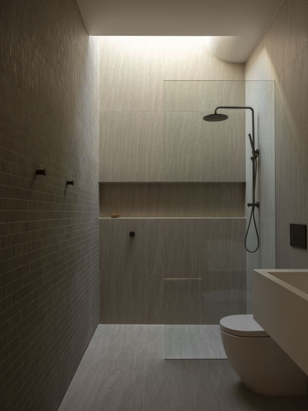 A compact, contemporary bathroom with a walk-in shower enclosed by a frameless glass screen. Large grey-beige stone-look tiles cover the floor and walls, while a dark small-format tiled wall contrasts with the minimalist white wall-hung vanity and rounded toilet. Black shower fittings, tapware, and flush plate add understated contrast. A skylight or high-level opening provides soft natural illumination.