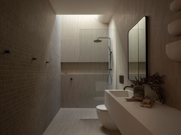 A narrow, sophisticated bathroom with a walk-in shower at the rear, wall-mounted shower fittings, a wall-hung toilet, and a long floating vanity with an integrated basin. The space features warm grey-beige textured wall and floor finishes, small-format tile on one wall, a large rounded-corner mirror, concealed lighting, and restrained natural styling. The atmosphere is dim, calm, and luxurious.