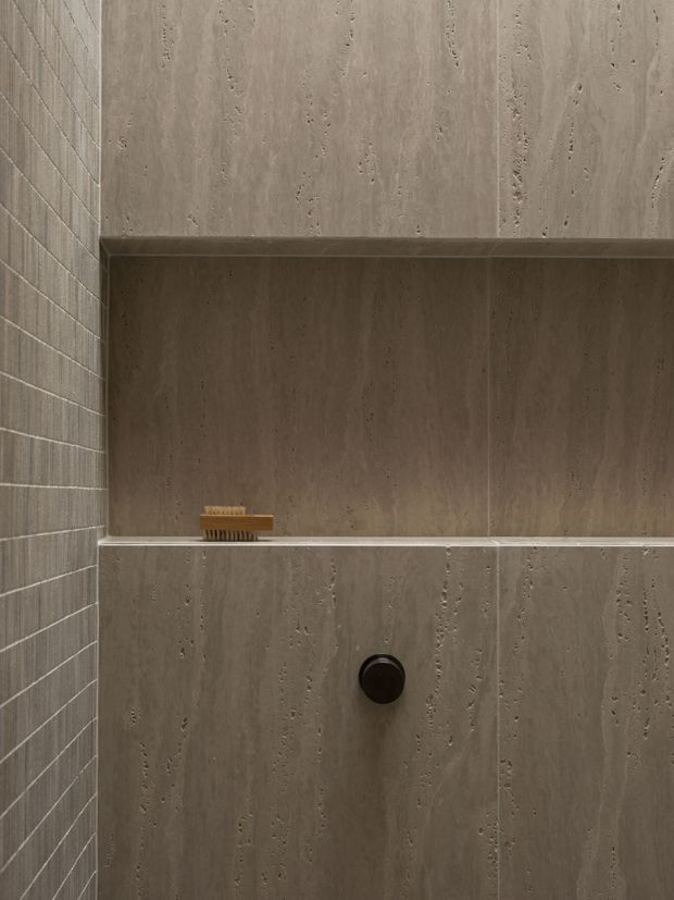 Close-up of a minimalist shower enclosure clad in taupe-grey stone-look slabs, with a recessed wall niche and integrated ledge. A small dark shower control is set into the lower wall, while the left wall features small-format grey tiles. The restrained material palette creates a refined, moody bathroom setting.
