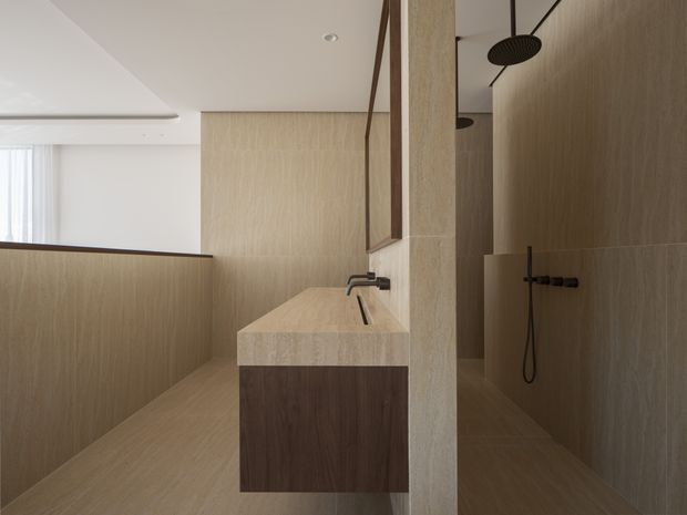 Minimalist contemporary bathroom with a floating dark timber vanity, integrated pale stone or porcelain basin, brushed dark metal tapware, and a walk-in shower. Warm wood-look wall and floor surfaces create a calm, cohesive interior, with a large mirror and partial view toward a bright adjacent space.